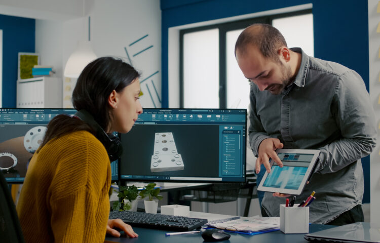 Industrial designer discussing with colleague holding tablet with in CAD program, designing 3D prototype of building. Woman engineer using pc with two monitors sharing ideas for construction mechanism